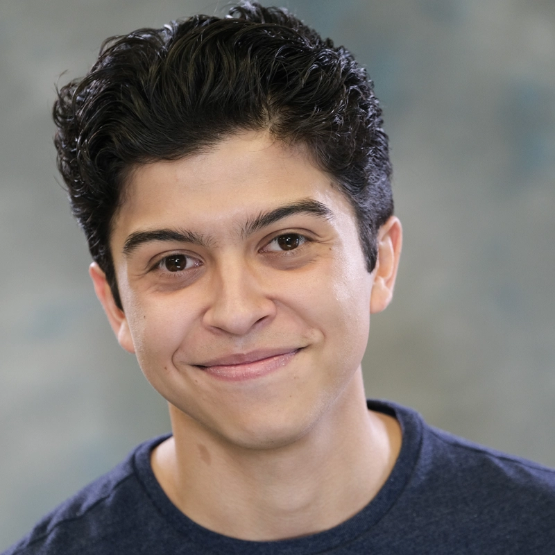 A young person with short, dark wavy hair smiles gently at the camera. Giovanny Diaz de Leon is wearing a dark blue crew-neck shirt, and the softly blurred gray and blue background keeps the focus on their friendly expression.