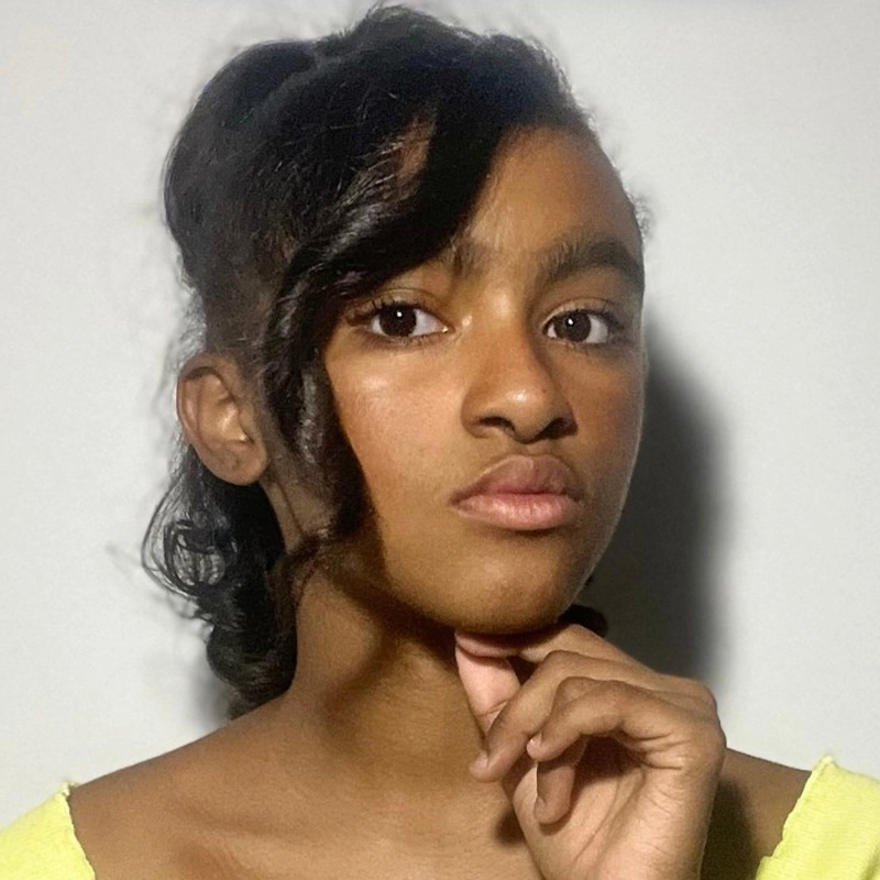 Shasteline Dilone, a young woman with medium brown skin and dark hair styled in loose waves, looks directly at the camera, resting her chin lightly on her hand. She wears a light yellow top against a plain, light background.