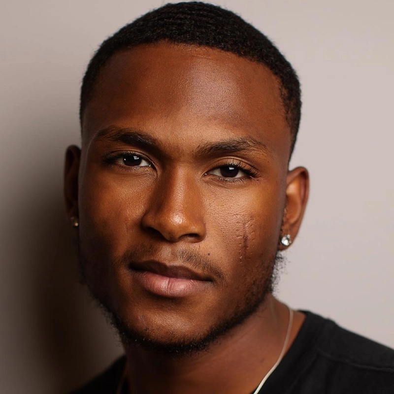 A young man with short black hair and trimmed facial hair looks slightly past the camera. He has a visible scar on his right cheek and wears stud earrings and a thin chain necklace. The background is plain and softly lit, highlighting his thoughtful expression.