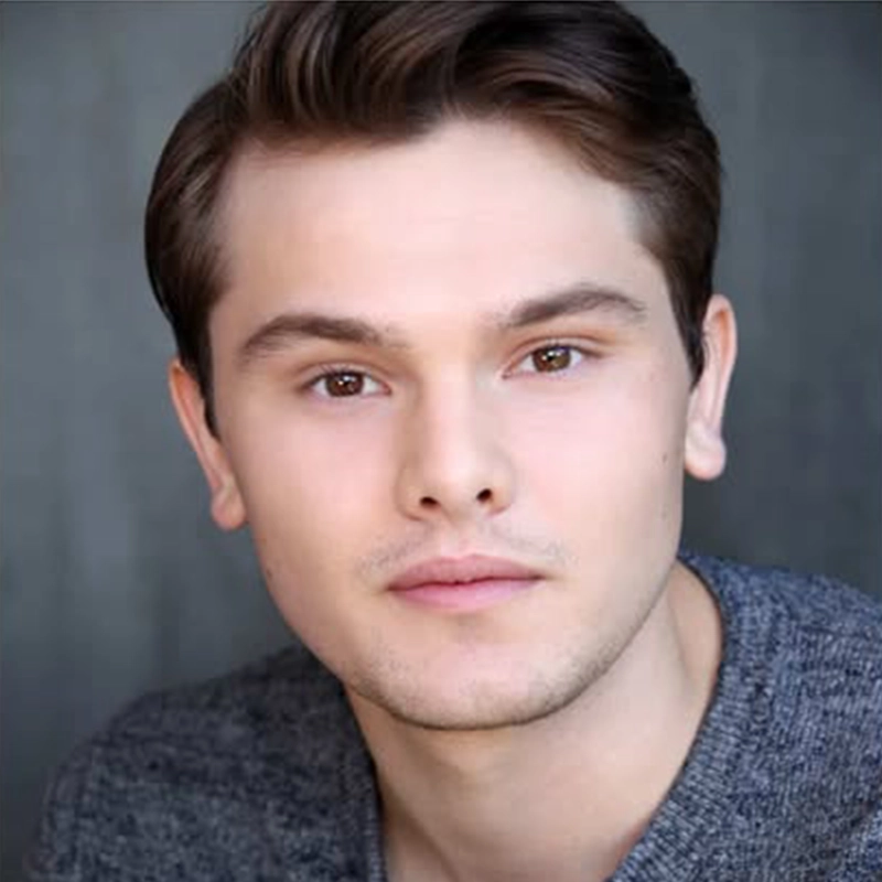 A young man with light skin, short brown hair, and brown eyes faces the camera with a neutral expression. He is wearing a gray textured sweater, and the background is softly blurred in shades of gray. The lighting is even and highlights his features.
