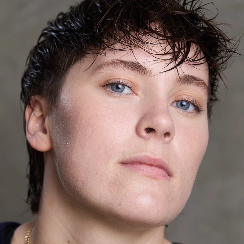 Close-up of Irene Emahiser with short, wet, dark hair, light skin, and blue eyes. They have a neutral expression and are looking slightly off-camera. The plain, out-of-focus background draws attention to Irene’s face.