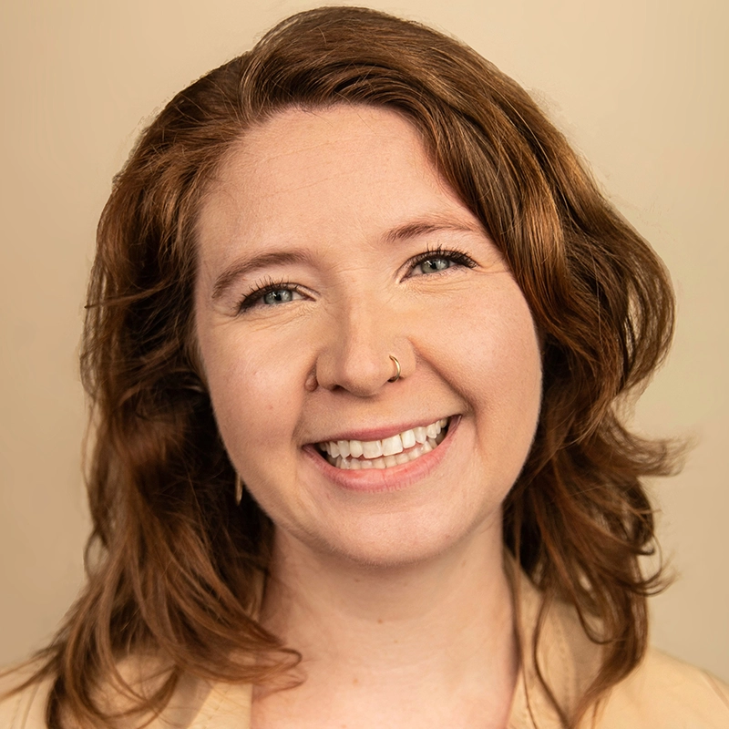 A woman with wavy, reddish-brown hair smiles warmly. She has fair skin, blue eyes, a nose ring, and wears light makeup. The background is a soft beige, and she is dressed in a tan top. The image conveys a friendly and approachable mood.