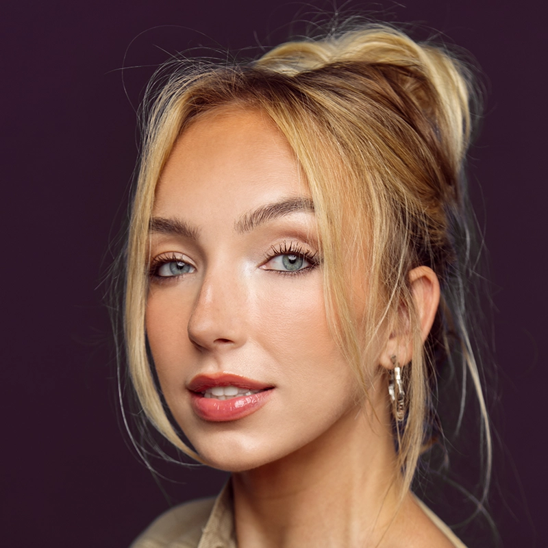 A young woman with fair skin and blue eyes looks at the camera. She has blonde hair styled in a loose updo with face-framing strands. She wears natural makeup and hoop earrings, set against a dark purple background.