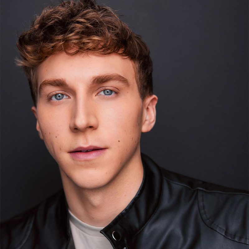 Mathew Fedorek, a young man with curly light brown hair and blue eyes, looks at the camera. He wears a black leather jacket over a light shirt against a plain dark gray background, with even lighting highlighting his face.