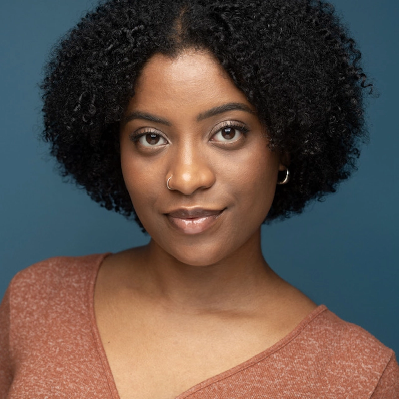 Jailyn Feliz, a young woman with short, curly black hair, smiles softly. She has a medium brown complexion, wears a rust-colored V-neck top, silver hoop earring, and nose ring. The background is solid blue.