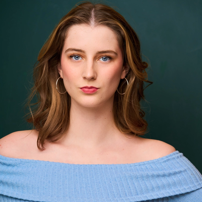 Cleo Fiedler, a young woman with wavy, shoulder-length brown hair and blue eyes, faces the camera against a dark green background. She wears a light blue off-the-shoulder top and large silver hoop earrings, her expression neutral.