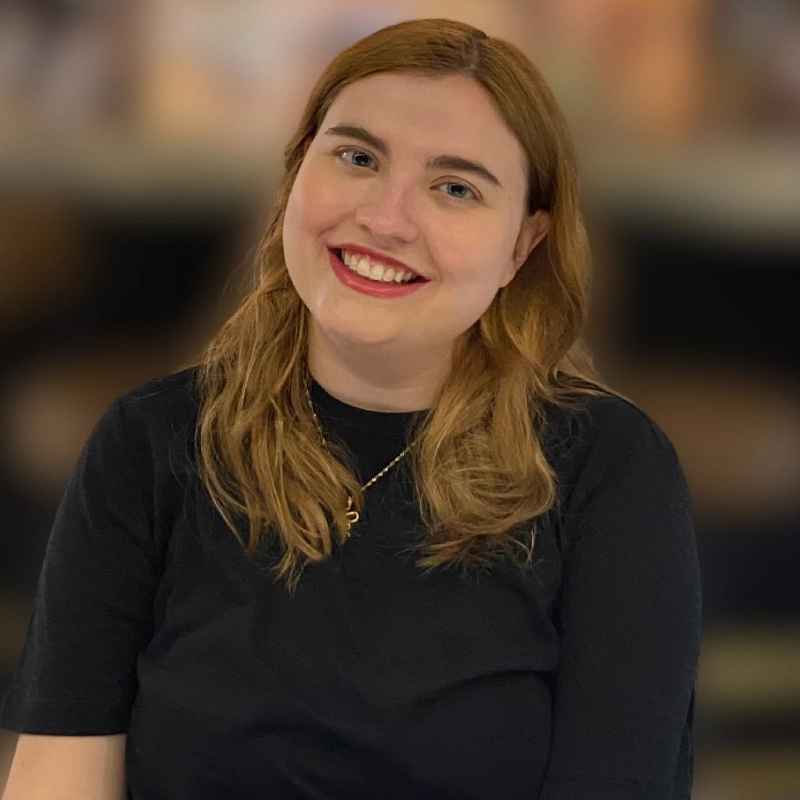 A young woman with fair skin and long, light brown hair smiles at the camera. She wears a black shirt and a gold pendant necklace. The background is softly blurred with warm tones, keeping the focus on her friendly expression.