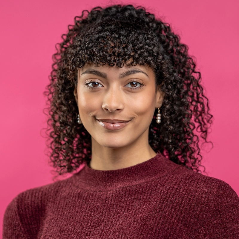 A woman with curly dark hair and bangs smiles softly. She wears pearl drop earrings and a maroon knit top. Alloria Frayser stands before a solid bright pink background, creating a vibrant contrast in this portrait-style photo.