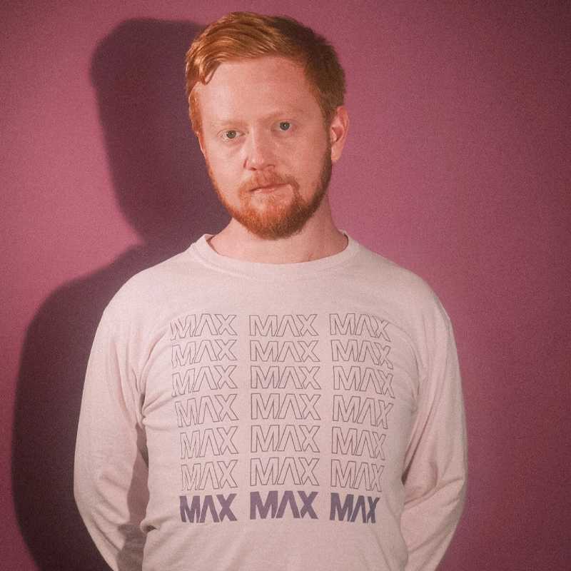 A man with fair skin and reddish hair and beard stands against a mauve background. He wears a light-colored long-sleeve shirt with “MAX” printed in purple, referencing Max Friedman. His expression is neutral, and his shadow is visible on the wall.