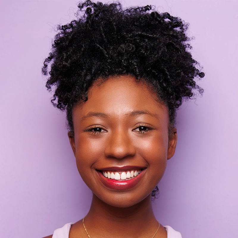 Isabel Marin Young, with dark curly hair in a high puff, smiles warmly. She wears natural makeup, red lipstick, and a lilac top. The solid light purple background adds to the cheerful and vibrant portrait.