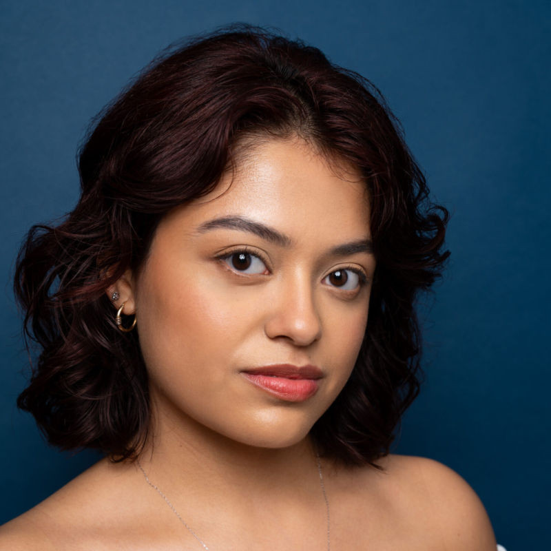 A young woman with wavy, shoulder-length dark brown hair poses against a solid blue background. She has light brown skin, natural makeup, and wears small hoop earrings. Her expression is calm and confident, looking directly at the camera.