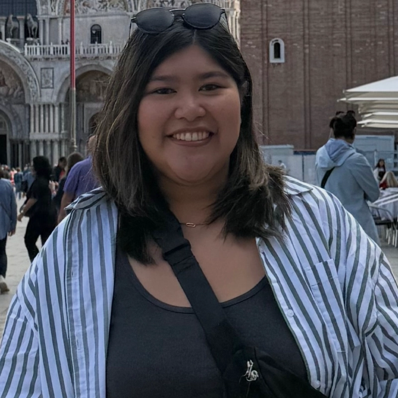 Kathleen Rose Gallardo, with straight dark hair, smiles at the camera. She wears sunglasses on her head, a striped button-up shirt over a dark tank top, and a crossbody bag. People walk near an ornate building with arches and columns behind her.