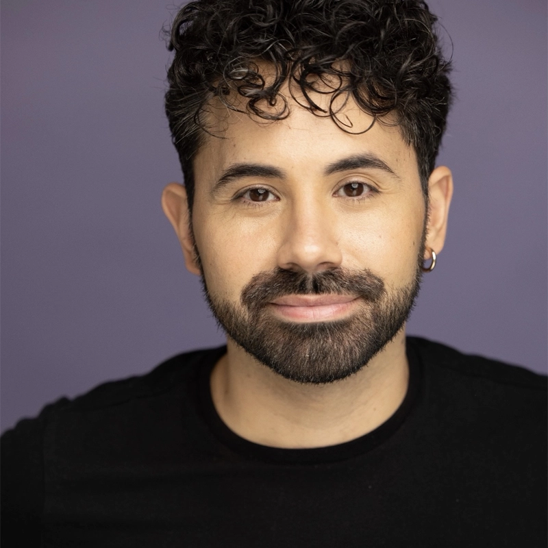 Omar Garibay, with curly dark hair, a trimmed beard, and mustache, smiles softly. He wears a small hoop earring in his left ear and a black shirt. The solid purple background and even lighting highlight his facial features.
