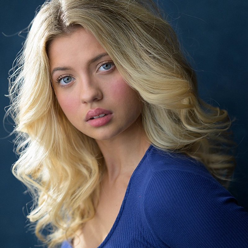A young woman, Katie Rose Garthe, with long, wavy blonde hair and blue eyes gazes softly at the camera. She wears a fitted, long-sleeve blue top and stands against a dark blue background. Her makeup is minimal with a hint of pink on her cheeks.