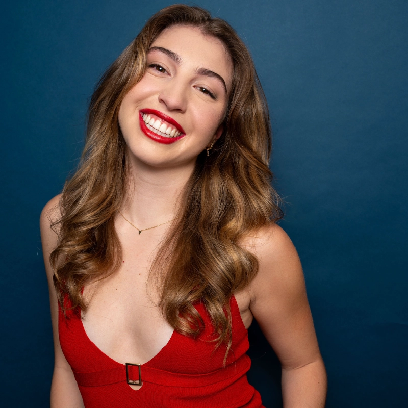Madeline Glave, a young woman with long wavy brown hair, smiles brightly against a dark blue background. She wears a sleeveless red dress with a deep neckline, a simple gold necklace, and bold red lipstick that matches her dress.