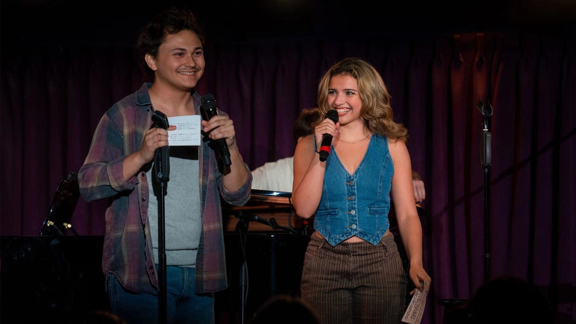 A young man in a plaid shirt and a young woman in a blue vest—Grant Sparr and Meg Gallo—smile on stage, each holding a microphone and note cards. They stand before a purple curtain, with a piano and microphone stands behind them at the New Year event.