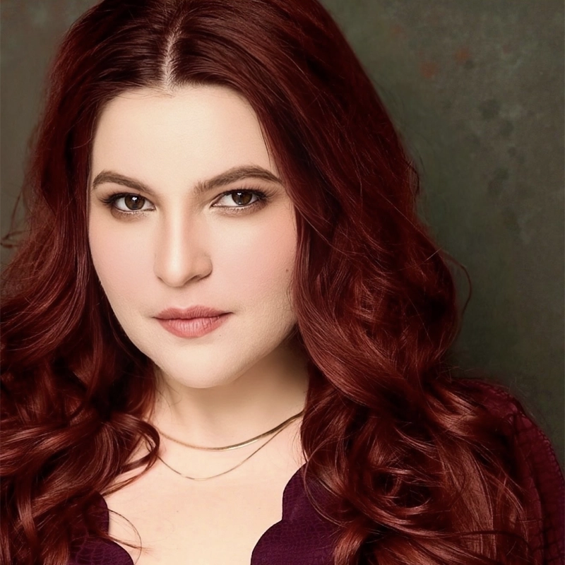 Gabrielle Gutierrez, with long, wavy dark red hair, poses against a textured dark background. She has fair skin, brown eyes, natural makeup, a burgundy top, and a delicate gold necklace, gazing confidently at the camera.