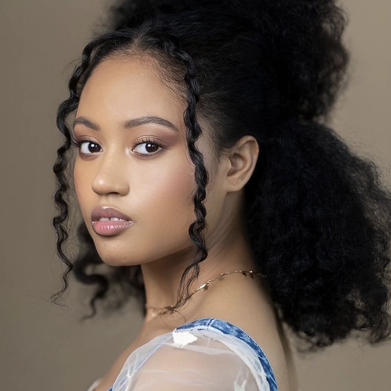 Lulu Oro Hamlett, a young woman with medium brown skin and voluminous, curly black hair, looks at the camera over her shoulder. She has defined eyebrows, natural makeup, and wears a sheer top with denim straps against a soft beige background.