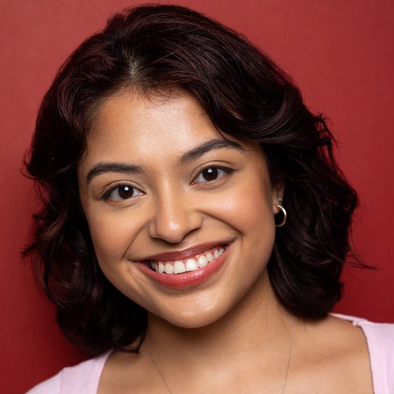 Gabriela Henriquez, a young woman with wavy, shoulder-length dark hair, smiles warmly at the camera. She wears small hoop earrings and a light pink top against a vibrant red background, with natural, subtle makeup.