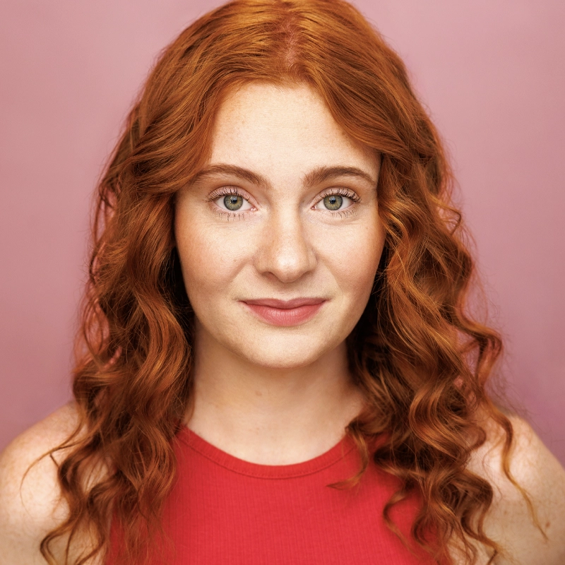 A young woman with long, curly red hair and fair skin smiles gently. Brynn Hofmeister wears a sleeveless red top and stands against a solid pink background, looking directly at the camera with green eyes and natural makeup.