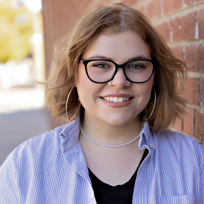 Payton Kelly Homer, a young woman with short, wavy light brown hair and glasses, smiles at the camera. She wears large hoop earrings, a double-strand pearl necklace, and a blue striped shirt over black, standing by a brick wall in soft daylight.