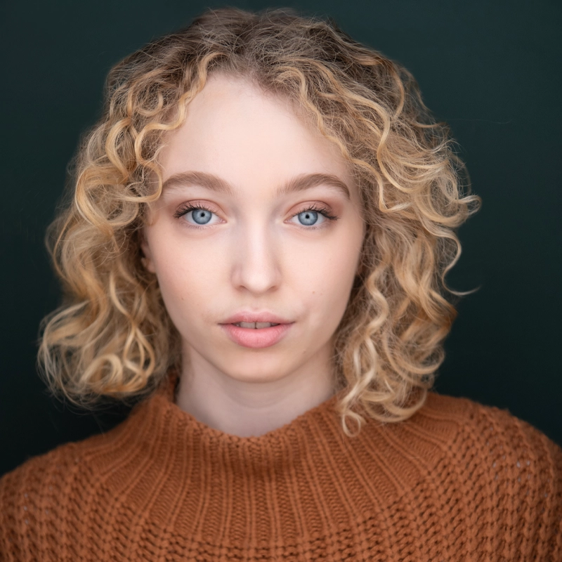 A young woman with light skin, blue eyes, and short, curly blonde hair looks directly at the camera. Emma Iannuzzi wears a textured brown sweater and neutral makeup. The dark green background contrasts with her sweater and hair.