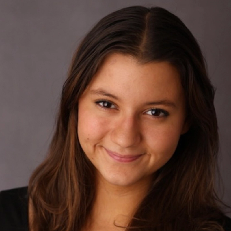Isabel Marin Young, with long brown hair and brown eyes, smiles gently at the camera. She wears a black top against a plain, soft gray background, her expression friendly and approachable.