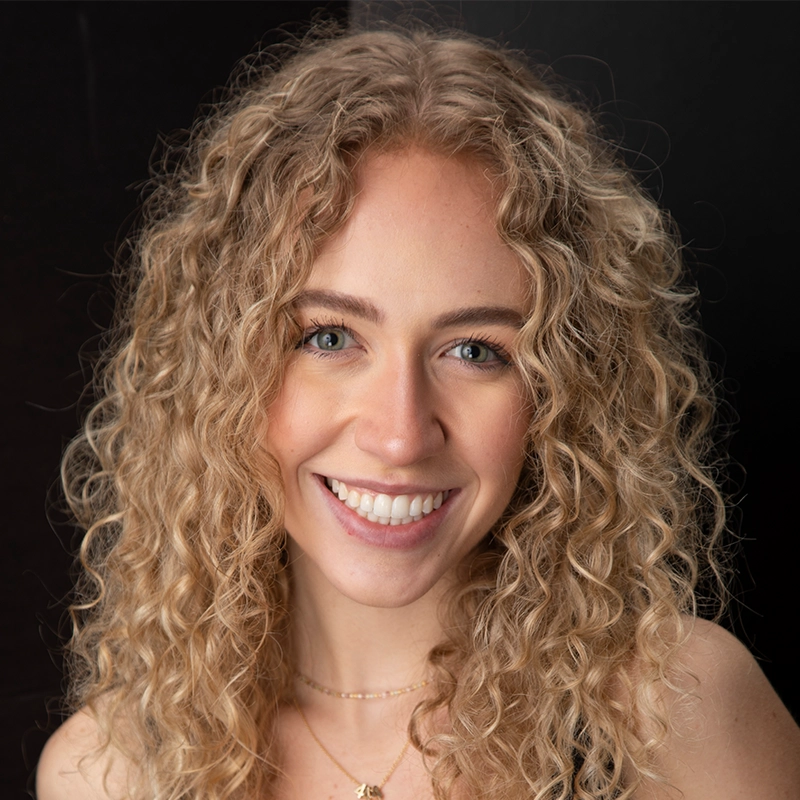 A young woman, Isabel Marin, with long, blonde, curly hair smiles warmly at the camera. She has light blue eyes and clear skin, wearing a necklace. The dark background makes her happy, relaxed expression stand out.