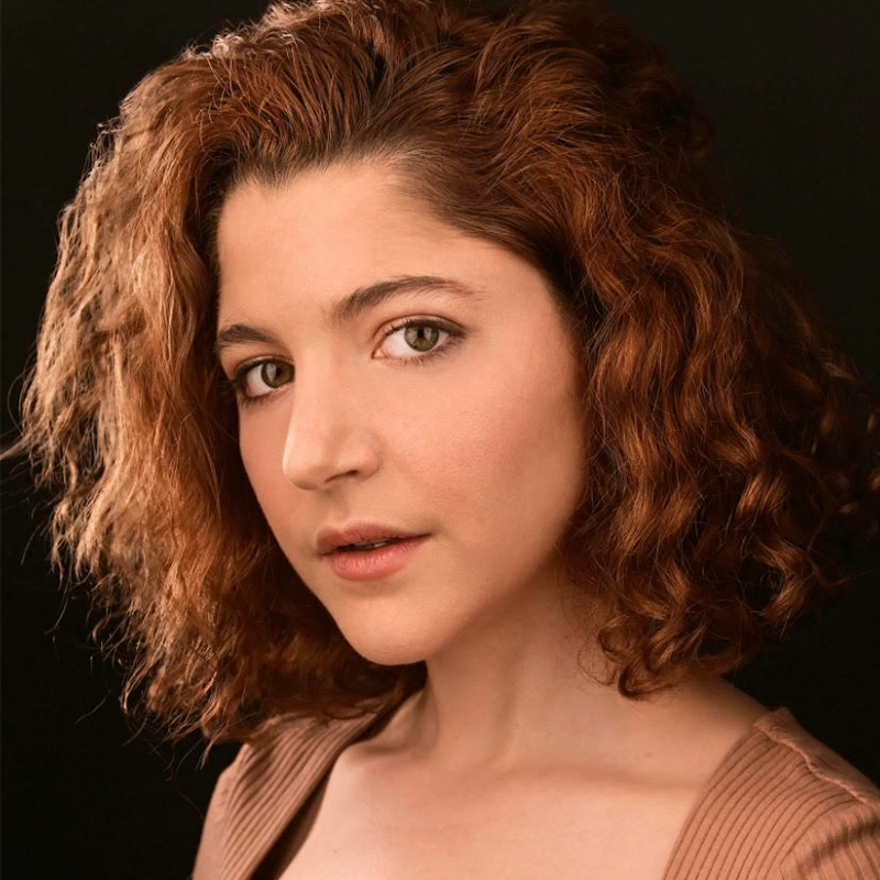 A woman with fair skin and shoulder-length, curly auburn hair looks at the camera with a neutral expression. She wears a light brown, ribbed top off one shoulder. The background is solid black, highlighting her features.