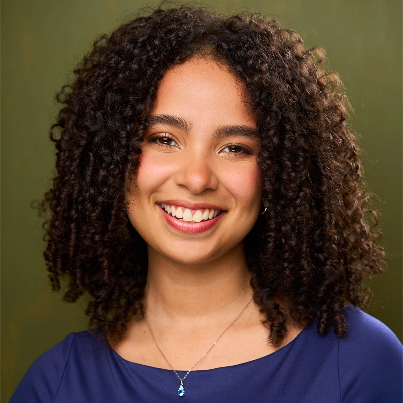 A young woman named Chanelle with medium skin tone, curly dark hair, and brown eyes smiles warmly at the camera. She is wearing a blue top and a delicate necklace with a small blue pendant. The background is an olive green color.