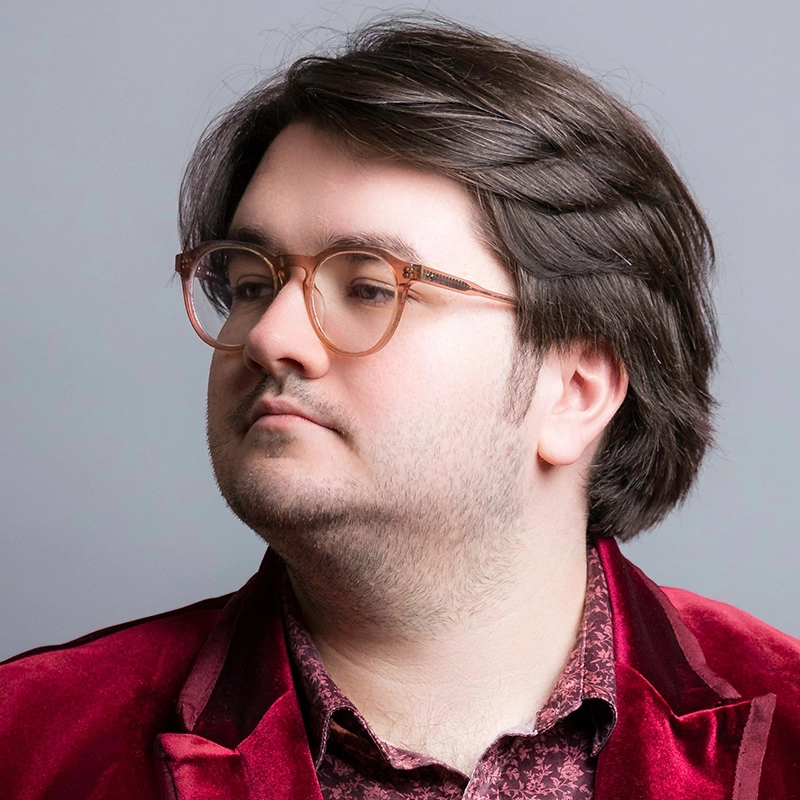 A young man with fair skin, straight dark brown hair, and a trimmed beard wears clear-framed glasses. He has a slight, neutral expression and looks to the left. He is dressed in a maroon velvet jacket over a patterned shirt against a gray background.