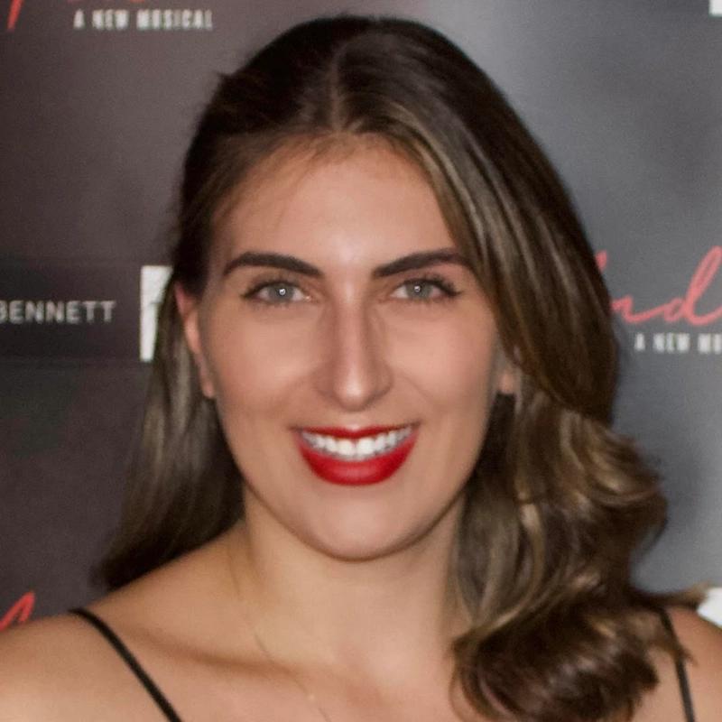 A woman with long brown hair and red lipstick smiles at the camera. She wears a black top with thin straps. The background features a dark event step-and-repeat banner with partially visible white and red text.
