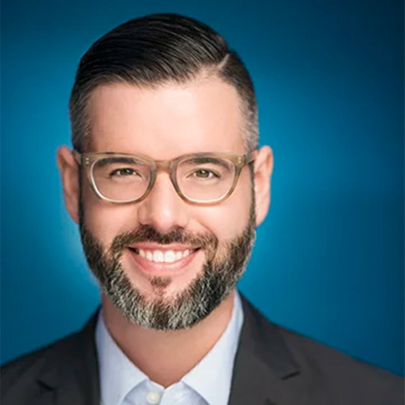 A man with dark hair and a trimmed beard, wearing clear-framed glasses, a dark blazer, and a light blue collared shirt, smiles against a gradient blue background—a style reminiscent of Samantha Kotok's professional portraits.