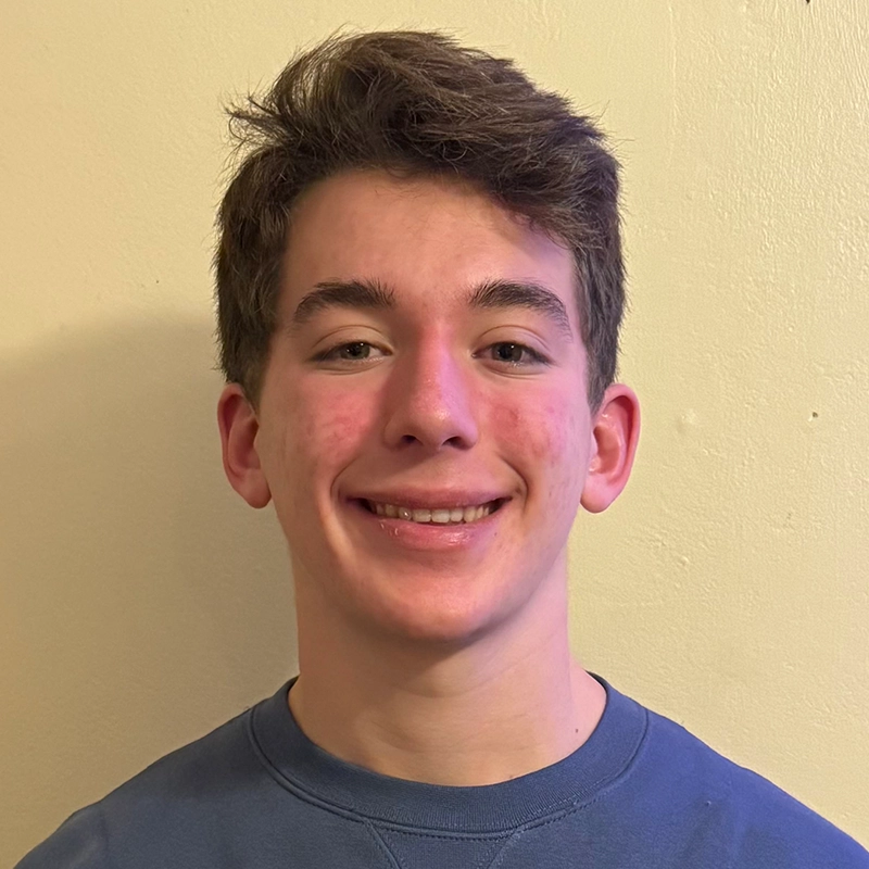 Isabel Marin Young, a young person with short brown hair and fair skin, smiles at the camera. They wear a blue crew-neck shirt and stand before a plain, light-colored wall, lit by soft, even lighting.