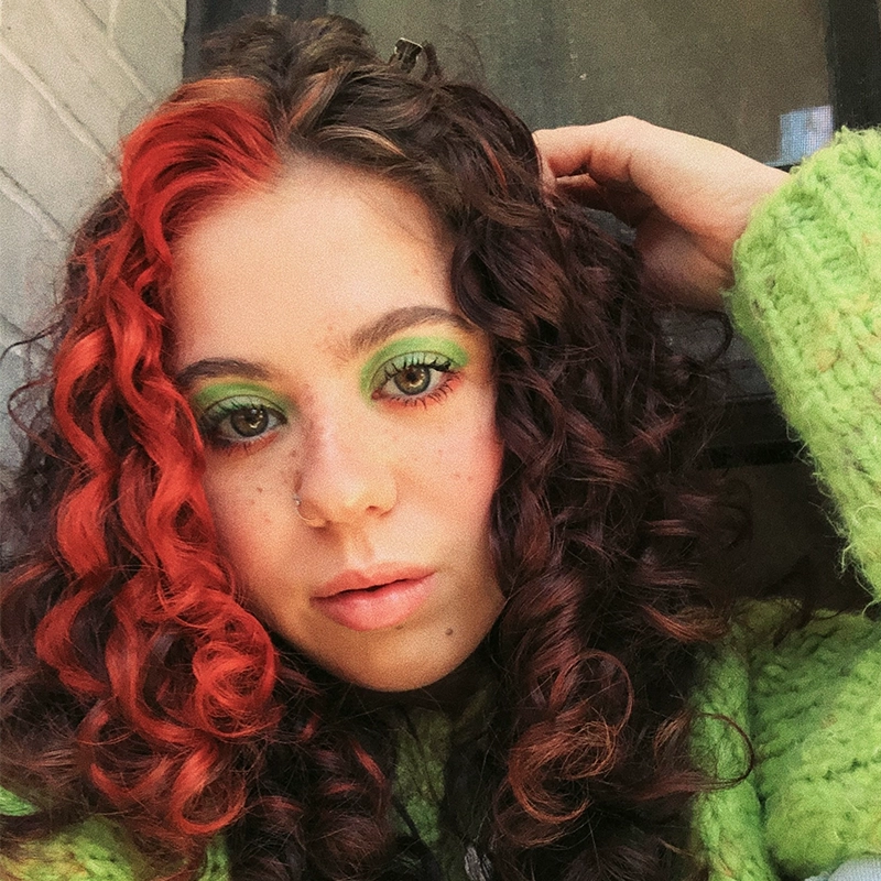 A young person with curly hair dyed black with a bright red section in front poses for a selfie. They wear bold green eye makeup and a fuzzy green sweater. One hand is raised to their head, and they have a nose ring and natural freckles.