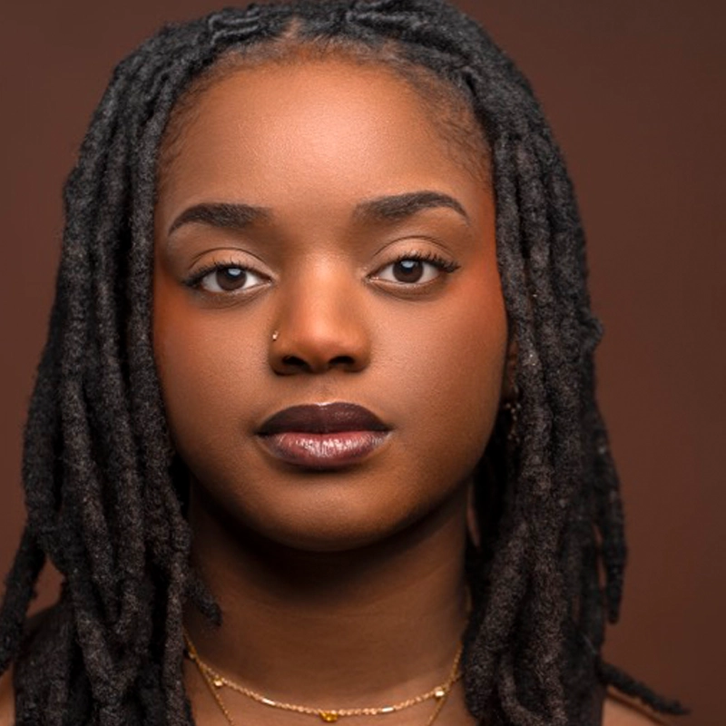 A woman with medium-dark skin, long black locs, and a nose piercing gazes confidently at the camera. She wears natural makeup, a neutral lip color, and layered gold necklaces. The background is a solid brown shade.