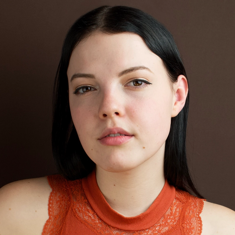 A young woman with fair skin and straight, dark hair is wearing an orange sleeveless top with lace details. She is looking directly at the camera with a neutral expression. The background is plain and brown.