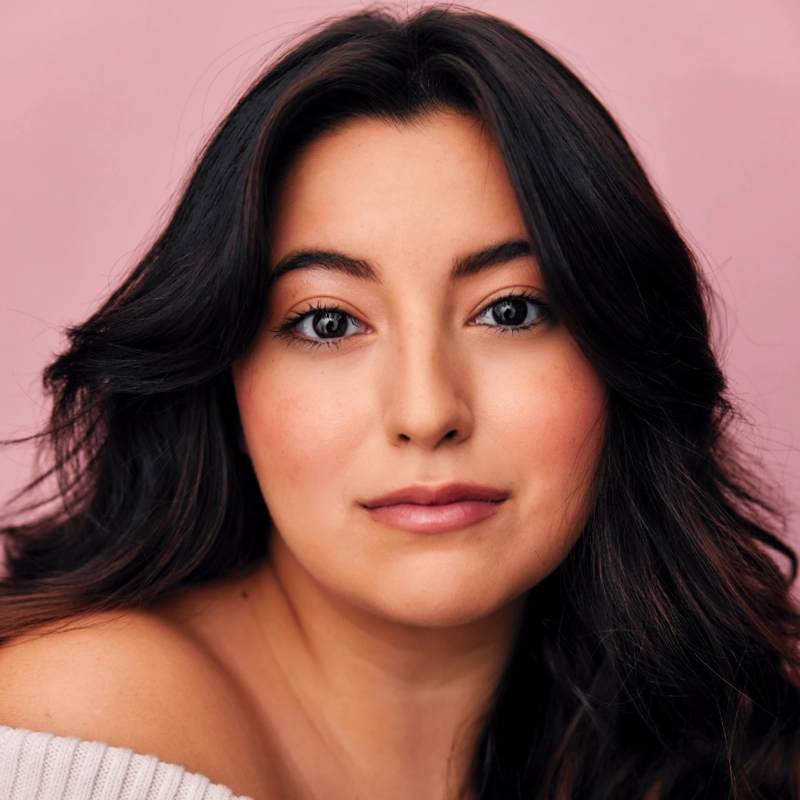 A young woman with long, wavy dark hair looks at the camera, her expression calm. Lucia Padilla Katz has smooth, light skin, natural makeup, and soft pink lips. She wears an off-shoulder white top against a solid, light pink background.