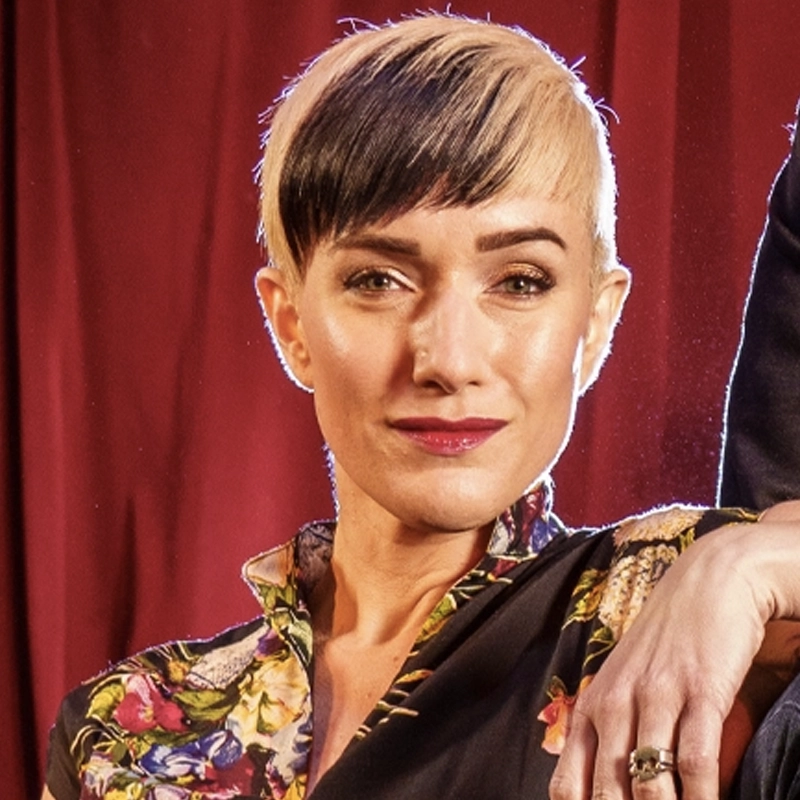 A confident person with short, asymmetrical blonde and black hair poses in front of red curtains. Styled like Kimberly Kaye, they wear a floral-patterned collared blouse and have a subtle smile, one arm resting casually on another person out of frame.