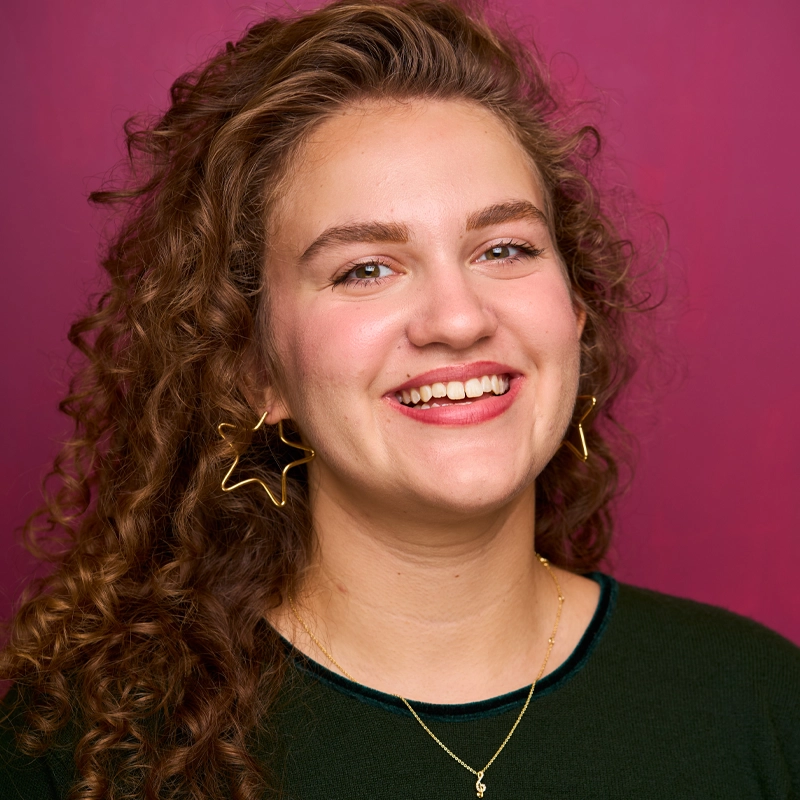 Natalie Kitchin, a young woman with long curly brown hair, smiles confidently in front of a magenta background. She wears a dark green top, gold star-shaped earrings, and a gold necklace, giving her natural makeup look an added cheerful glow.