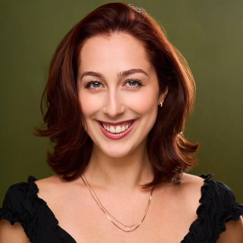 Zola Kneeland, with shoulder-length red hair, smiles at the camera in a black top and two gold necklaces. The smooth olive green background and lighting highlight her clear skin and bright eyes.