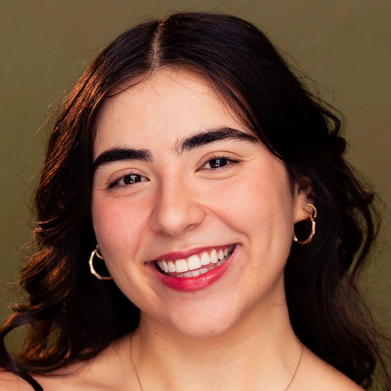 A young woman with wavy dark hair smiles warmly at the camera. She wears gold hoop earrings and natural makeup, and her skin tone is light. The background is a soft olive green, creating a calm and inviting portrait atmosphere.