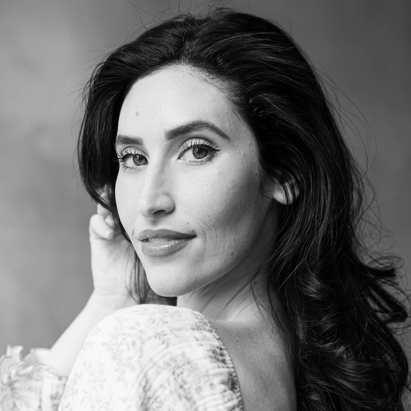 Black-and-white portrait of Lauren Paley with long, wavy dark hair, looking over her shoulder at the camera. She has defined eyebrows, long eyelashes, and wears a patterned off-the-shoulder top against a softly blurred background.
