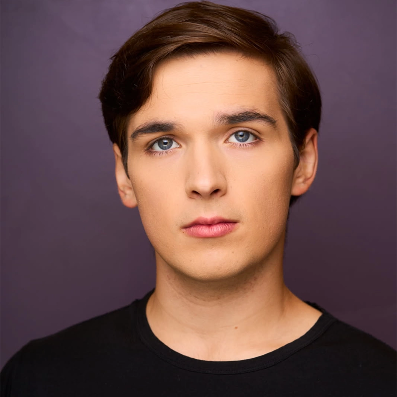 Javen Levesque, a young person with fair skin, short brown hair, and blue eyes, looks directly at the camera with a neutral expression. They are wearing a black crewneck shirt and standing against a plain purple background.