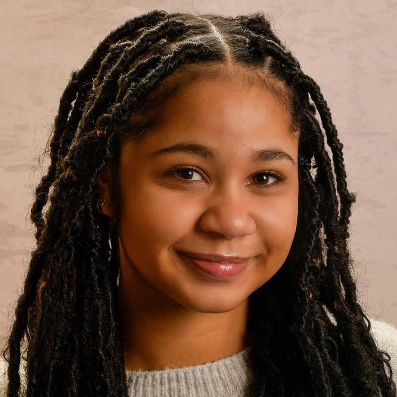 A young woman with long, dark, twisted braids smiles softly at the camera. Vita Augugliaro has warm brown skin, dark eyes, and wears a light grey sweater. The pale, textured background creates a calm and inviting portrait atmosphere.