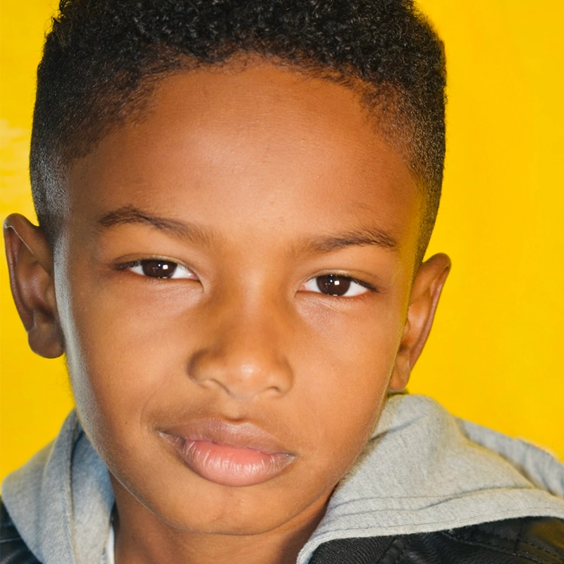 A close-up of Lincoln Alejandro Collier, a young boy with short curly hair and medium brown skin, looking directly at the camera. He wears a gray hoodie under a black jacket, his neutral expression set against a bright yellow background.