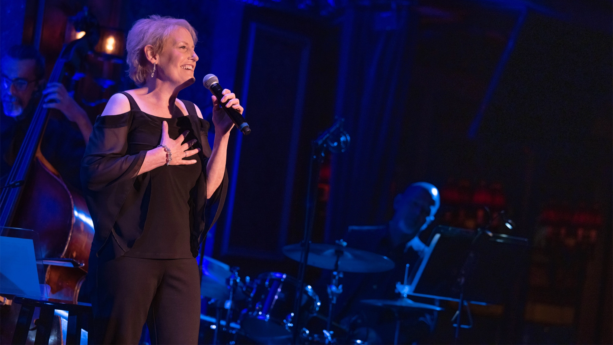 Liz Callaway - Show Art A woman with short blonde hair, dressed in a black cold-shoulder blouse and pants, sings into a microphone on stage during a Sondheim birthday celebration. She holds one hand to her chest as the bassist and drummer play under warm blue and purple lighting.