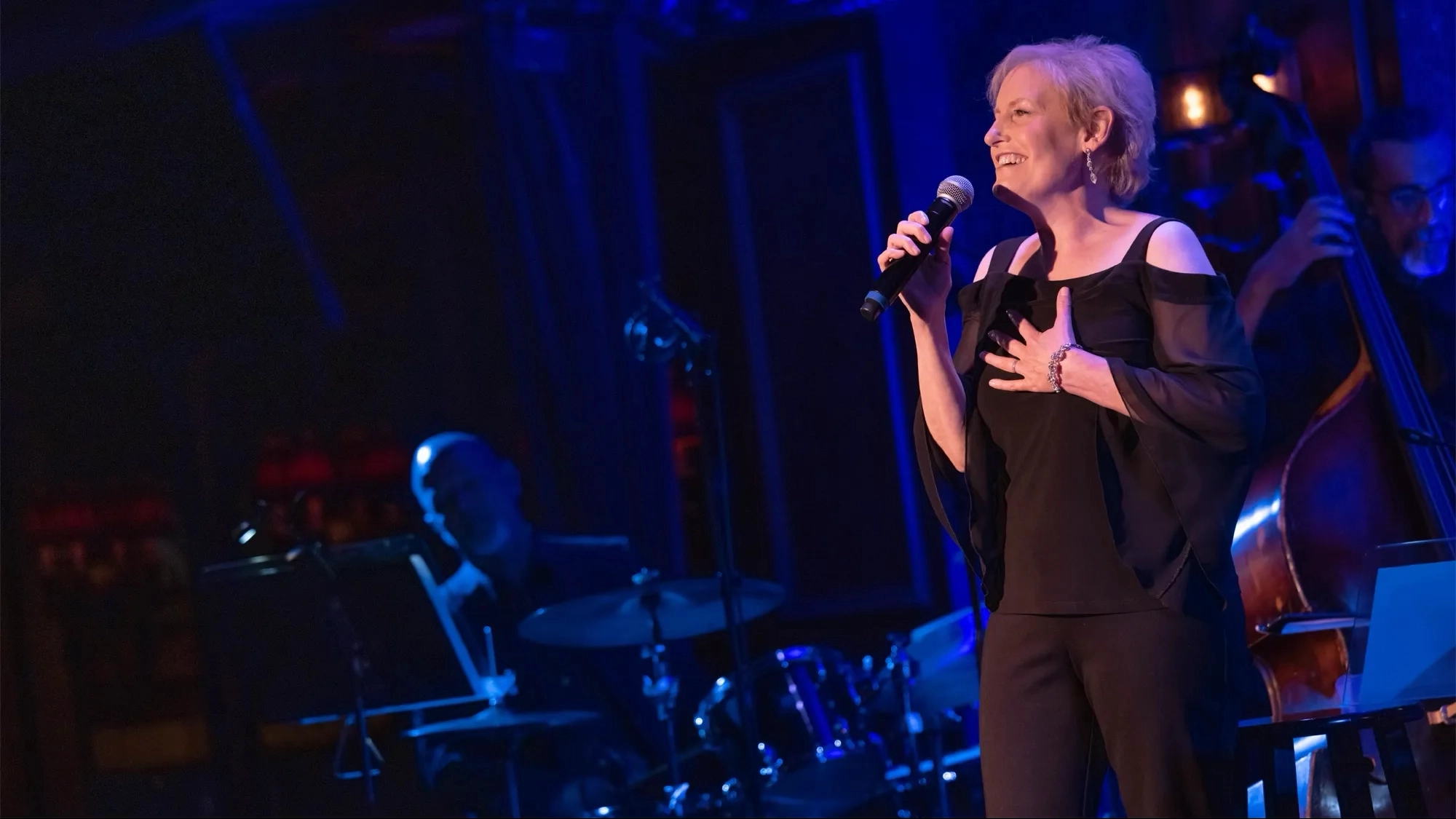 A woman with short blonde hair, dressed in a black outfit with cutout shoulders, holds a microphone and sings onstage. She smiles and rests a hand on her chest as blue stage lights shine—celebrating Sondheim’s birthday alongside her band.
