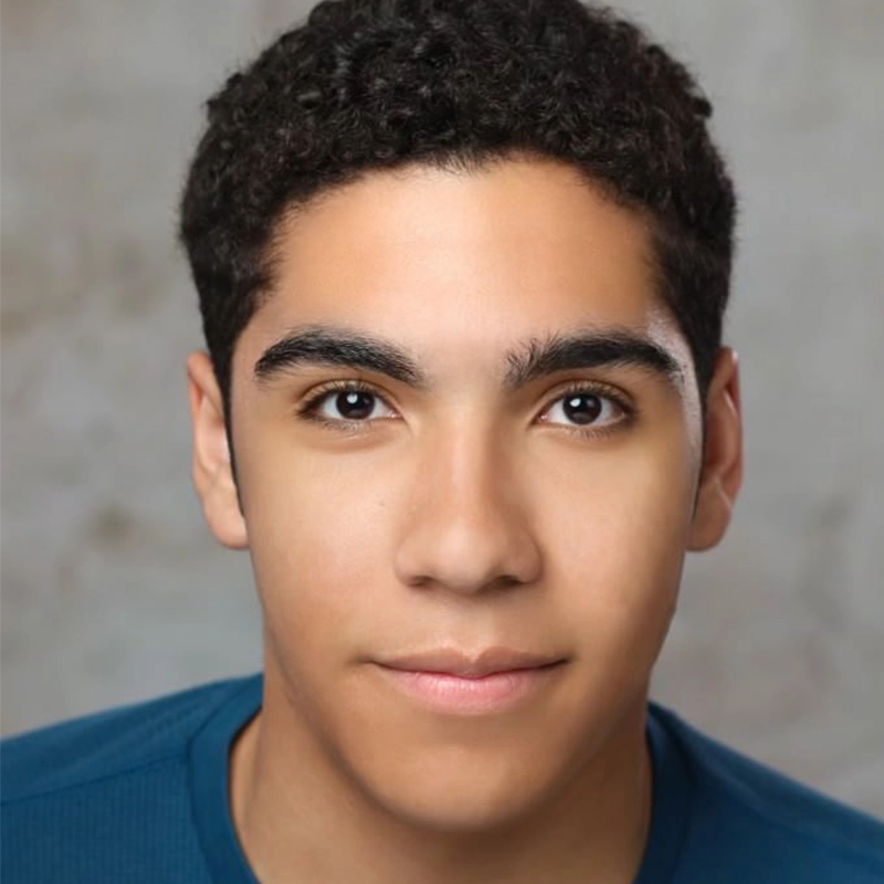 A young person with short, curly dark hair and smooth skin looks directly at the camera. With dark eyes and defined eyebrows, Isabel Marin Young wears a blue shirt against a softly blurred, light-colored background.