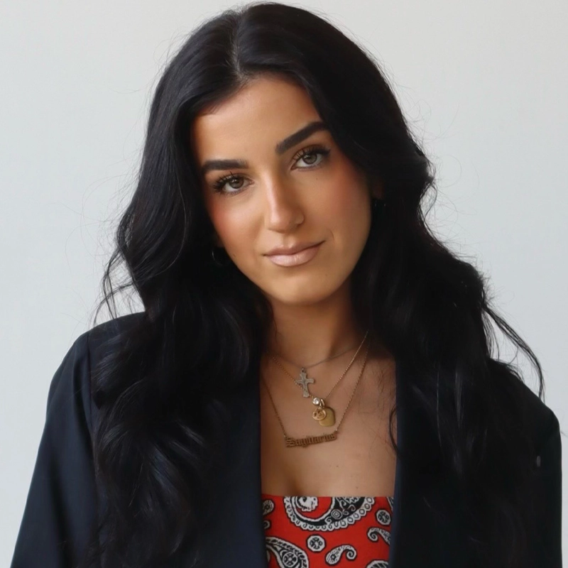Sofia Luetich, with long, wavy black hair, poses against a plain white background. She wears a black jacket over a red and white paisley-patterned top and layered gold necklaces. Her makeup is natural with strong brows.